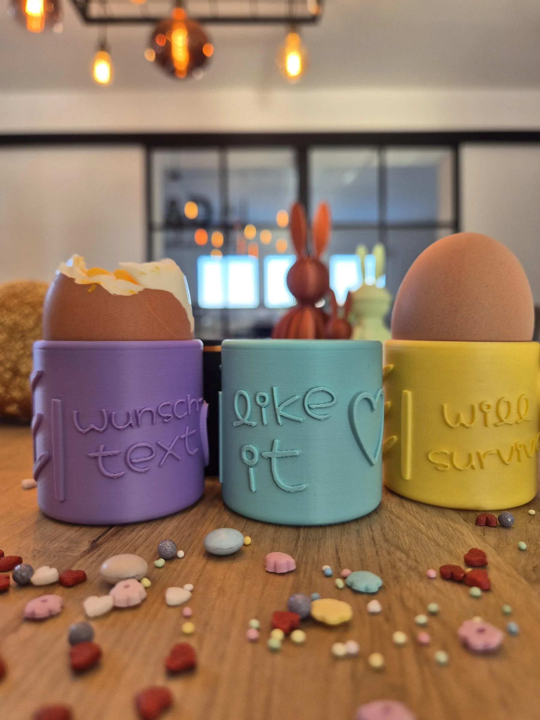 Drei Eierbecher in Lavendel, Mint und Gelb mit „Wunschtext“, „EI like it“ und „EI will survive“ auf dem Frühstückstisch